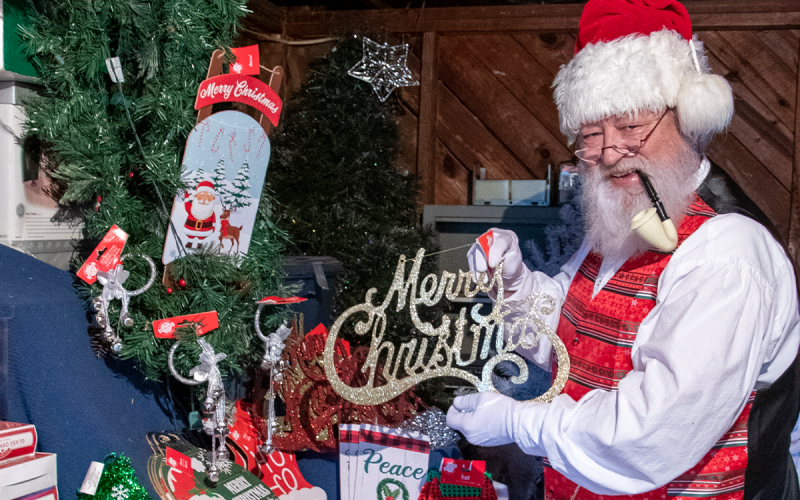 The Sophisticated Santa, also known as Bob Williams, stands in a traditional Santa pose, complete with pipe and “Merry Christmas” sign. Williams, who lives in Cornelia, uses proceeds from his Santa appearances to fund his “Shop Santa’s Garage,” where families who need them can get Christmas decorations, including trees, ornaments and stocking stuffers at no charge. KIMBERLY BROWN/Staff