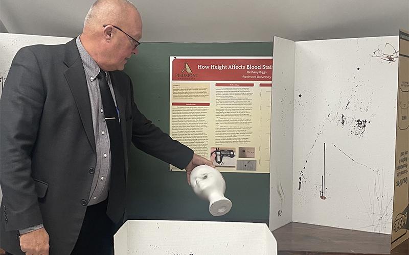 Piedmont University Vice President of Academic Affairs Bruce Willis holds an example of a gunshot wound to the head next to students’ assignments on trajectory of blood stains. JULIANNE AKERS/Staff