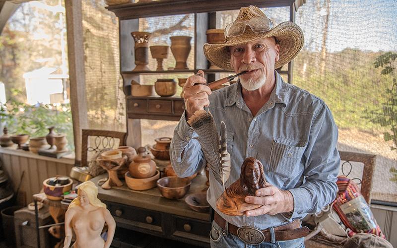 Kenneth Adams stands next to a collection of completed pieces, holding a carved Native American man with feathers and a handmade pipe from his collection of past works. He carves and turns many pieces of wooden art each month, ranging from projects that have lasted years to spontaneous projects that come to his mind on three-day weekends. ZACH TAYLOR/Staff
