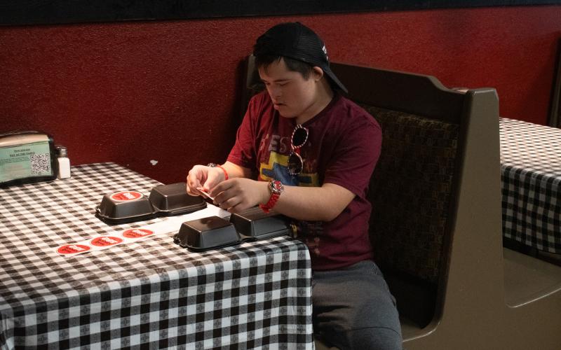 Nathan Dooley sits under a light at one of the booths in the Wolf Creek BBQ diner, concentrating on the task of putting stickers on to-go boxes.  HANNAH CAUDELL/Staff