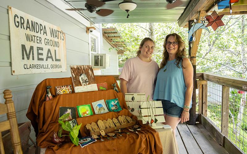 Erin Witkowski and Aubry Evans pose on the porch of Mark of the Potter as Witkowski shows and sells her glass work and Evans displays her paintings for the North Georgia Arts Tour.