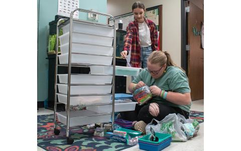 Holly Samsel (left) hands a pair of scissors to Emmalee Turgeon (right) as she tries to open packaged makeup bags for her new hygiene cart, which is part of her 4-H Leadership in Action project. (Hannah Caudell/Staff)
