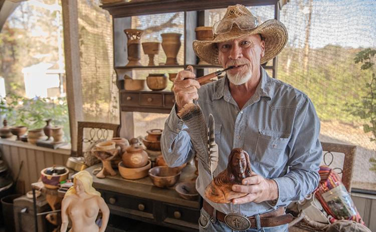 Kenneth Adams stands next to a collection of completed pieces, holding a carved Native American man with feathers and a handmade pipe from his collection of past works. He carves and turns many pieces of wooden art each month, ranging from projects that have lasted years to spontaneous projects that come to his mind on three-day weekends. ZACH TAYLOR/Staff