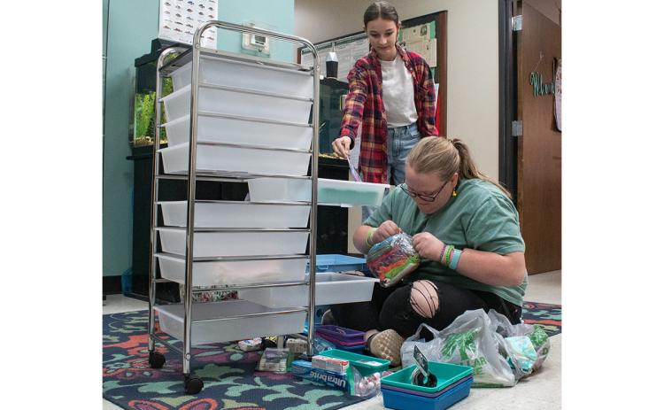 Holly Samsel (left) hands a pair of scissors to Emmalee Turgeon (right) as she tries to open packaged makeup bags for her new hygiene cart, which is part of her 4-H Leadership in Action project. (Hannah Caudell/Staff)